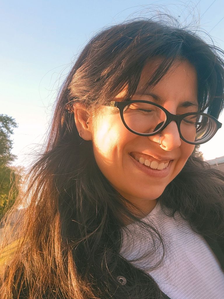A person with long dark hair and glasses smiling against a clear sky.